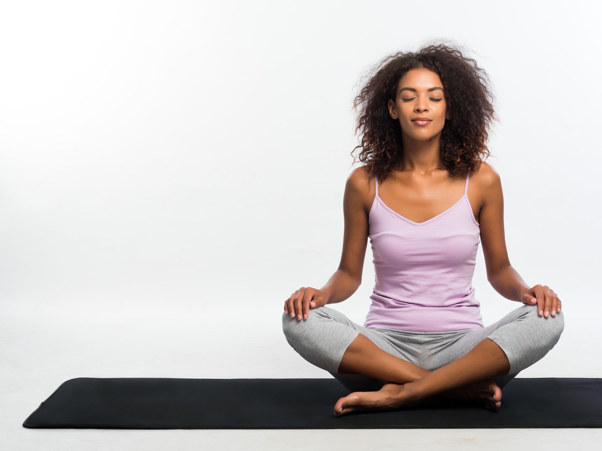 Happy african american woman in comfortable sports wear meditating on mat over white wall background. Black girl concentrated on yoga practice.