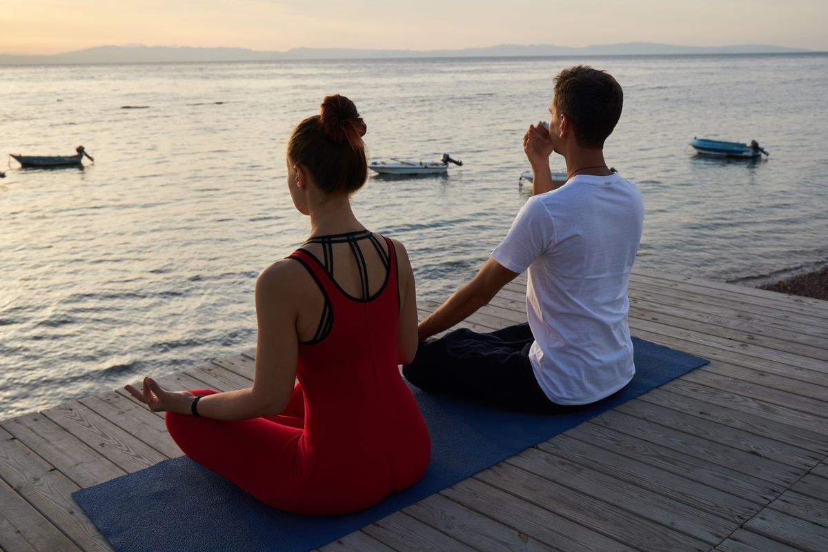 Male and female sitting on wooden terrace by the sea and practicing meditation and pranayama